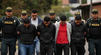 Siete hombres con caras pixeladas, algunos con chaquetas y jeans, son escoltados por policías colombianos en uniforme y chalecos con la palabra "POLICIA".