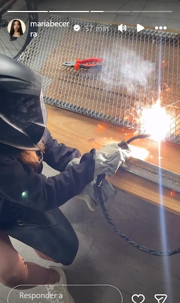 Una persona con casco de soldar y guantes blancos está soldando metal, produciendo chispas brillantes y humo sobre una superficie de madera y una malla metálica