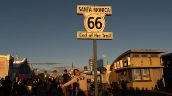 Una persona con una sudadera gris con el logo "GAP" posa con los brazos abiertos bajo el letrero "SANTA MONICA 66 End of the Trail" al atardecer en el muelle