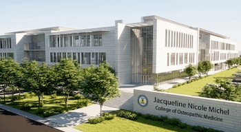 Exterior de un moderno edificio con fachada de piedra clara y grandes ventanales, rodeado de áreas verdes con árboles y un letrero identificativo