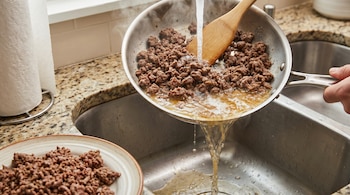 Carne picada cocida en una sartén, escurrida con agua caliente sobre un fregadero de cocina. La grasa y el agua caen. Hay una espátula y un plato con carne magra.