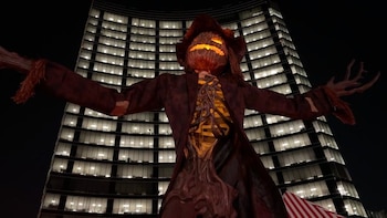 Figura de espantapájaros con cabeza de calabaza iluminada y brazos abiertos frente a un edificio iluminado de noche y una carpa rayada
