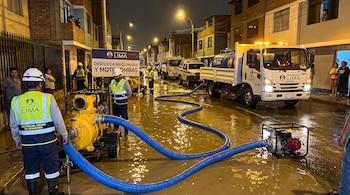 Calle inundada de noche con operarios, motobombas amarillas, mangueras azules, vehículos de la Municipalidad de Lima y un cartel.