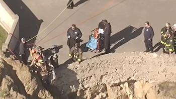 Vista aérea de bomberos y personal de rescate en un acantilado, atendiendo a una persona con arnés y cuerdas, con una camilla cerca en el suelo