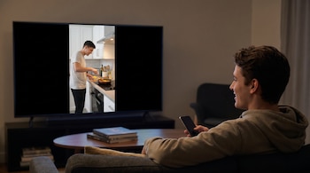 Un hombre en un sofá viendo un video vertical de cocina en un televisor grande en una sala oscura. Sostiene un smartphone en su mano derecha.