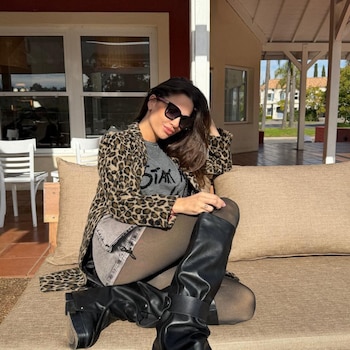 Ivana Figueiras posa sentada en un sofá exterior, luciendo una chaqueta con estampado de leopardo, camiseta gris, shorts y botas altas negras, en un ambiente soleado.