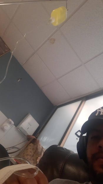 Un hombre con gorra y auriculares recibe una infusión intravenosa de líquido amarillo en un entorno de clínica, vista en ángulo