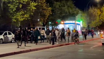 Vista nocturna de una calle con un grupo de peatones en el centro, autos estacionados a la izquierda y edificios iluminados al fondo