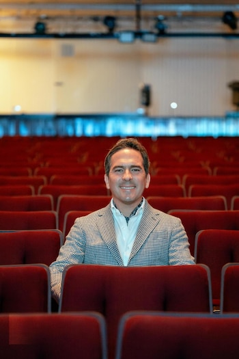 Hombre sonriente de camisa blanca y saco a cuadros sentado en una butaca roja de teatro vacía, con otras filas de asientos detrás
