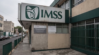Letrero grande con logo IMSS, letras IMSS, y dos letreros institucionales menores en la fachada de un edificio con ventanas y una reja verde en la calle.