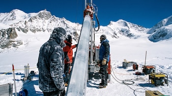 Tres glaciólogos en un glaciar operan una perforadora que extrae una columna de hielo. Hay montañas nevadas y cielo azul de fondo, y equipo científico.