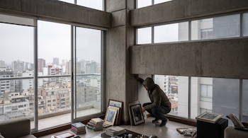 Un hombre agachado junto a una columna de concreto en un apartamento. Grandes ventanales muestran edificios de una ciudad