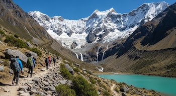 Grupo de excursionistas con mochilas camina por un sendero de piedras junto a una laguna turquesa. Al fondo, montañas nevadas y cascadas bajo cielo azul.
