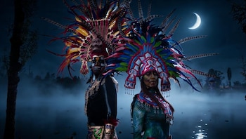 Dos figuras con tocados de plumas multicolores y maquillaje facial están de pie en un lago con niebla bajo la luna creciente