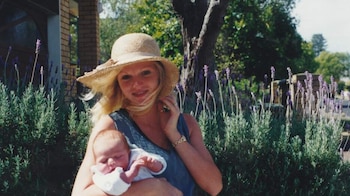 Una mujer rubia con sombrero de paja sostiene a un bebé dormido en brazos. Detrás hay plantas de lavanda, un árbol y una construcción de ladrillo