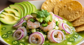 Aguachile de camarón servido en un plato blanco con rebanadas de aguacate, rodajas de cebolla morada, chiles serranos y hojas de cilantro fresco, con tostadas al fondo.