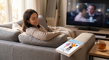 Una mujer joven con suéter beige se sienta en un sofá gris, sonriendo y mirando la televisión, con el libro "Romper el hielo" sobre el apoyabrazos.