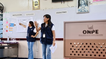 Dos mujeres con chalecos azules con las siglas ONPE están en un aula junto a carteles informativos y una caja electoral.