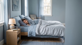 Una mujer duerme de costado en una cama con ropa azul, junto a una mesa de noche de madera con una lámpara encendida y un libro.