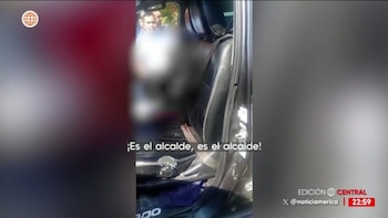 Captura de pantalla de un video que muestra el interior de un vehículo con un asiento de cuero oscuro y una figura borrosa. Dos hombres observan desde el exterior