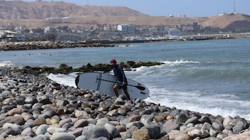 Un hombre con gorra y camiseta oscura transporta una tabla de paddle surf a la orilla de una playa de piedras frente al mar y cerros