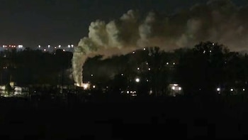 Una columna de humo espeso asciende en medio de la noche sobre luces urbanas distantes y vegetación oscura