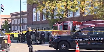 Un agente policial coloca una cinta de precaución frente a un vehículo de policía y un camión de bomberos ante un edificio de ladrillo