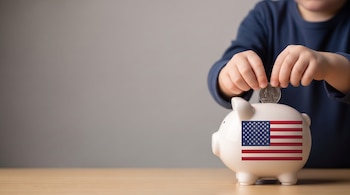 Manos de un niño que depositan una moneda estadounidense en una alcancía con forma de cerdito. La alcancía muestra la bandera de Estados Unidos.