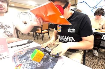 Hombre con los ojos vendados y una camiseta negra prepara sus manos sobre un temporizador junto a un cubo Rubik de 3x3 sobre un tapete