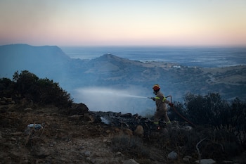 Bombero con uniforme ignífugo, casco y máscara rocía agua en terreno quemado. Al fondo, colinas, mar y cielo al amanecer o atardecer