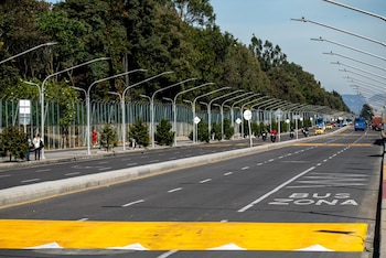 Carretera pavimentada con la marca BUS ZONA, paso peatonal amarillo, postes de luz, árboles, peatones, motos y autobuses al fondo