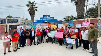 Un grupo de adultos protesta frente a un colegio con carteles. La fachada del colegio es azul con el nombre "República de Francia". Hay palmeras y un cielo nublado