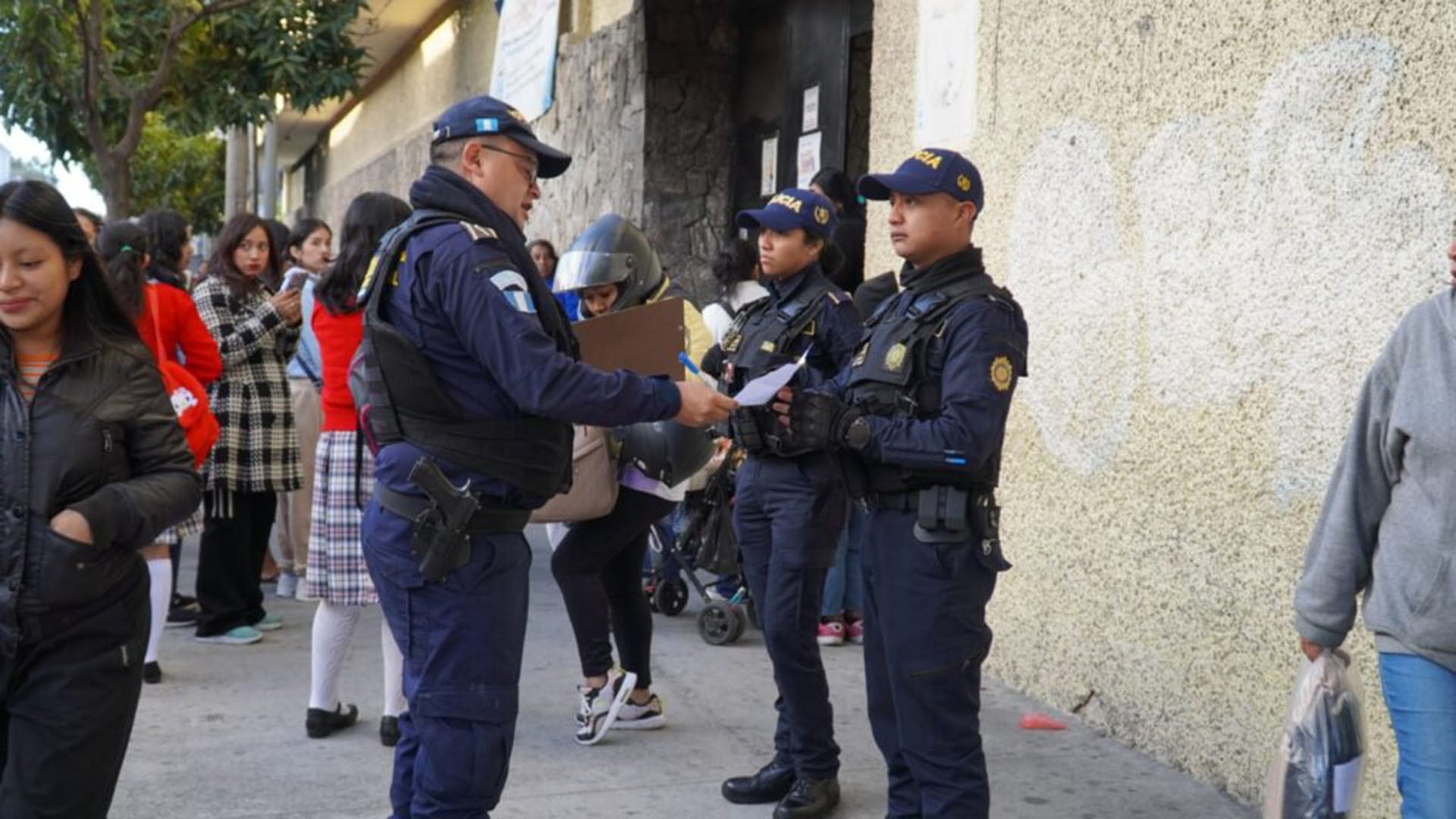 Agentes de la Policía Nacional Civil de Guatemala (PNCdeGuatemala) mantienen una presencia activa para garantizar la seguridad en los alrededores de una escuela guatemalteca, donde se observan estudiantes y peatones. (PNCdeGuatemala)