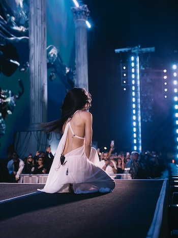 Mujer de espaldas con vestido blanco arrodillada en la pasarela de un escenario iluminado frente a la multitud en un concierto