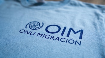 Primer plano de una camiseta de tela azul claro con el logo azul marino de OIM ONU Migración claramente visible en la parte frontal.