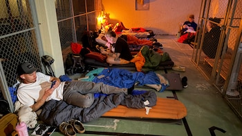 Varias personas descansan en sacos de dormir sobre colchonetas en el suelo de un espacio con cerramientos de malla metálica