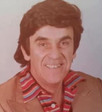 Primer plano de un hombre sonriente de cabello oscuro y abundante, viste camisa a rayas horizontales rojas, blancas y azules con saco marrón
