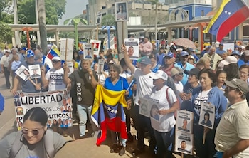 Multitud de personas marchan por una calle soleada sosteniendo banderas de Venezuela y pancartas con fotos de individuos, algunos con el texto 'Libertad'