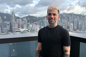 Un hombre con cabello rubio y barba posa al aire libre. Tiene tatuajes en el cuello y los brazos. Al fondo, un horizonte urbano con montañas y rascacielos