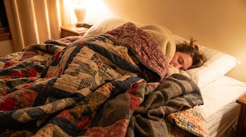 Persona durmiendo en una cama con almohada, cubierta por un edredón de retazos y múltiples mantas. Una lámpara de mesa ilumina la mesita de noche.