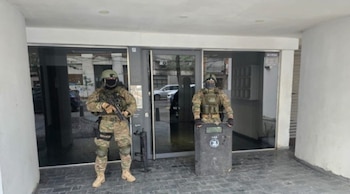 Dos agentes de seguridad con uniformes de camuflaje, cascos y máscaras, uno con un rifle y otro con un escudo táctico, posan frente a un edificio