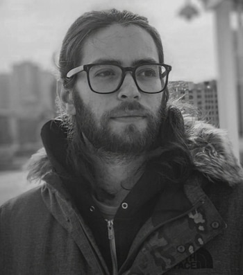 Retrato en blanco y negro de un hombre con gafas, barba, cabello largo. Lleva una chaqueta con capucha y un forro de piel sintética. Fondo urbano borroso