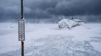 Termómetro de poste oxidado marcando ochenta grados Celsius bajo cero, una cabaña cubierta de nieve, y un extenso terreno helado bajo un cielo nublado y ventoso.