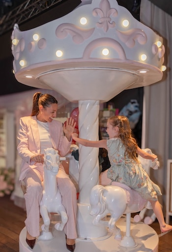 Una mujer y una niña ríen en un carrusel. El carrusel tiene caballos blancos, detalles rosados y luces. La mujer usa un traje rosa, la niña un vestido estampado