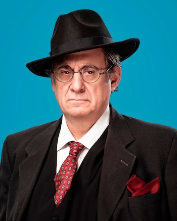 Un hombre con sombrero negro, gafas redondas, saco oscuro, camisa blanca, corbata roja con patrón y pañuelo rojo, sobre un fondo azul