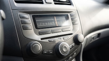 Panel de control de un automóvil con una radio de pantalla analógica con dial vertical, botones rectangulares y tres perillas giratorias.