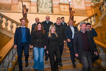 Un grupo de doce personas, hombres y mujeres, baja una escalera de madera. Paredes de mármol decoradas con estatuas y murales con 'PRO PATRIA' al fondo