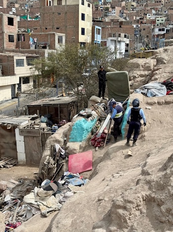 Vista aérea de tres hombres uniformados en una ladera rocosa llena de escombros y estructuras improvisadas, con edificios residenciales al fondo