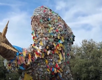 Escultura de gran tamaño elaborada con botellas, bolsas de plástico de colores y otros desechos, frente a un cielo azul con nubes y árboles
