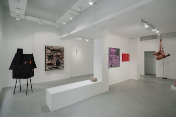 Interior de una galería de arte con paredes blancas y suelo gris, mostrando varias obras: una casa negra en patas, un cuadro texturizado, dos lienzos gráficos, y una escultura suspendida